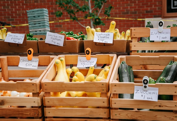 A farmer's market display featuring a variety of vegetables in wooden crates. There are signs indicating the types and prices, such as shishito peppers, zucchini, candy onions, and summer squash. The background consists of a red brick wall and green foliage.