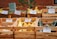 A farmer's market display featuring a variety of vegetables in wooden crates. There are signs indicating the types and prices, such as shishito peppers, zucchini, candy onions, and summer squash. The background consists of a red brick wall and green foliage.