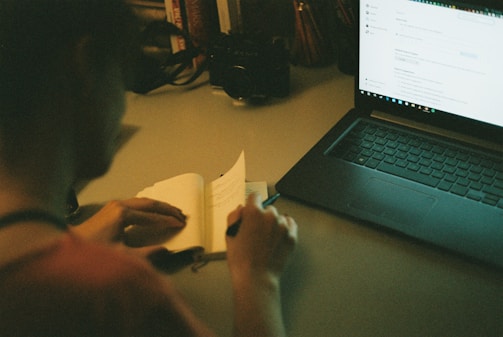 A person is sitting at a desk, writing in a notebook with a pen. A laptop is open on the desk, displaying a webpage. The scene is dimly lit, suggesting an evening or late-night work environment. Books and a camera are visible on the desk in the background.