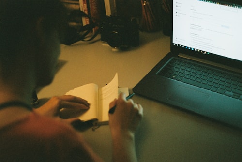 A person is sitting at a desk, writing in a notebook with a pen. A laptop is open on the desk, displaying a webpage. The scene is dimly lit, suggesting an evening or late-night work environment. Books and a camera are visible on the desk in the background.