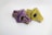 Two intricately designed face masks are laying side by side. One mask features a vibrant purple background with yellow and black paisley patterns, while the other displays a bright yellow backdrop with similar designs in purple and black. Both masks have black filters on the sides.