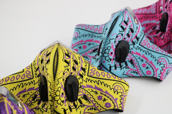 Three patterned face masks are displayed, each with distinct designs and vibrant colors. The masks feature intricate paisley patterns in shades of yellow with purple, blue with pink, and pink with black. Each mask is equipped with a black ventilation valve.