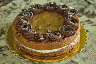 A round cookie cake adorned with chocolate frosting and small blue and white sprinkles sits on a gold platter. The cake has a layer of white cream sandwiched in the middle and a textured surface with chocolate chips visible.