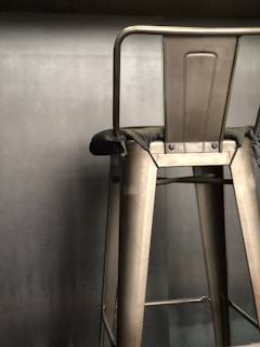 A metallic bar stool with a high backrest is positioned against a plain, muted background. The stool features strong, industrial lines and a slightly worn fabric covering its seat.