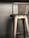 A metallic bar stool with a high backrest is positioned against a plain, muted background. The stool features strong, industrial lines and a slightly worn fabric covering its seat.