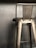 A metallic bar stool with a high backrest is positioned against a plain, muted background. The stool features strong, industrial lines and a slightly worn fabric covering its seat.