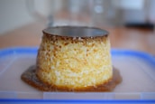 A creamy flan dessert sits on a clear plastic surface, topped with a layer of caramel. The dessert features a smooth texture and a rich golden-brown hue, with caramel sauce pooling at its base. Background elements are out of focus, drawing attention to the flan.