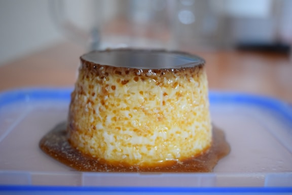A creamy flan dessert sits on a clear plastic surface, topped with a layer of caramel. The dessert features a smooth texture and a rich golden-brown hue, with caramel sauce pooling at its base. Background elements are out of focus, drawing attention to the flan.