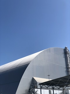 A large industrial structure with a curved roof, possibly a warehouse or a plant facility, is framed against a clear blue sky. A tall chimney or exhaust pipe is visible on one side, and metal framework or scaffolding can be seen in the foreground.