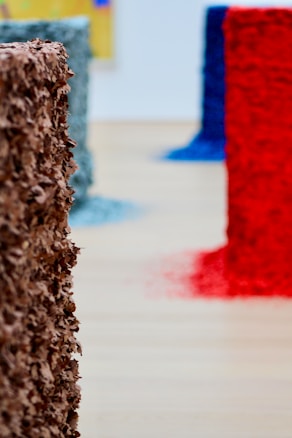 Several cylindrical sculptures, adorned with textured layers of different colors, are positioned on a wooden floor. The sculptures are primarily colored in earthy brown, vibrant red, deep blue, and muted teal, each with loose debris spreading around their bases.