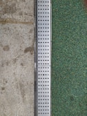 A close-up view of a surface transition featuring a concrete slab on the left and a textured green surface on the right. Separating the two areas is a metal strip with a series of evenly spaced circular holes, likely serving as drainage.