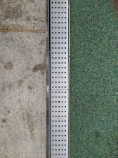A close-up view of a surface transition featuring a concrete slab on the left and a textured green surface on the right. Separating the two areas is a metal strip with a series of evenly spaced circular holes, likely serving as drainage.