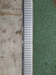 A close-up view of a surface transition featuring a concrete slab on the left and a textured green surface on the right. Separating the two areas is a metal strip with a series of evenly spaced circular holes, likely serving as drainage.
