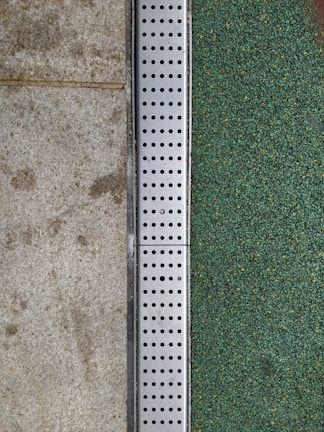 A close-up view of a surface transition featuring a concrete slab on the left and a textured green surface on the right. Separating the two areas is a metal strip with a series of evenly spaced circular holes, likely serving as drainage.