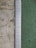 A close-up view of a surface transition featuring a concrete slab on the left and a textured green surface on the right. Separating the two areas is a metal strip with a series of evenly spaced circular holes, likely serving as drainage.