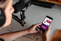 A person wearing headphones is seated at a desk with a microphone setup and a keyboard. They are holding a smartphone displaying a colorful audio recording app interface, with options for starting and managing recordings.