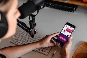A person wearing headphones is seated at a desk with a microphone setup and a keyboard. They are holding a smartphone displaying a colorful audio recording app interface, with options for starting and managing recordings.