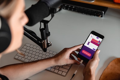 A person wearing headphones is seated at a desk with a microphone setup and a keyboard. They are holding a smartphone displaying a colorful audio recording app interface, with options for starting and managing recordings.