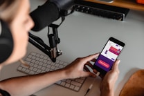 A person wearing headphones is seated at a desk with a microphone setup and a keyboard. They are holding a smartphone displaying a colorful audio recording app interface, with options for starting and managing recordings.