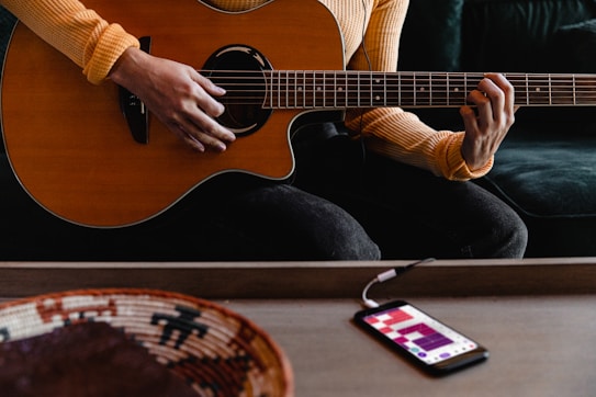 A person wearing a striped long-sleeve shirt playing an acoustic guitar. The guitar is held in a seated position with focus on the person's hands strumming and fretting. In the foreground, a smartphone is connected to a cable, displaying an application with a grid-like pattern. Next to the phone is a woven basket with geometric designs.