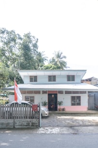 A two-story house with a sloped roof, featuring a fence in front and a car parked in the driveway. A large red and white flag is displayed outside. The house is painted in light colors, with green and pink accents. Surrounding the house are lush trees and plants, indicating a tropical setting.