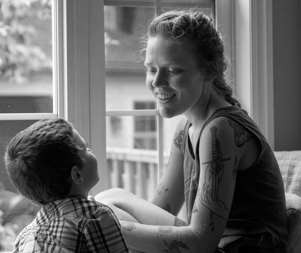 grayscale photo of woman in tank top sitting beside window