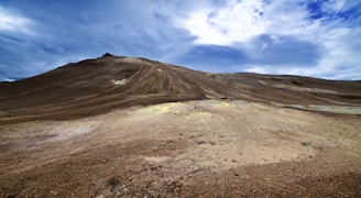 A barren, expansive rocky landscape with a gently sloping hill under a dynamic blue sky. The ground is covered with rough, brown earth and small patches of yellowish soil, indicating mineral deposits.