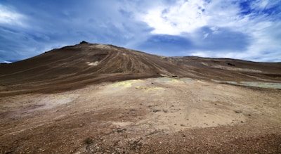 A barren, expansive rocky landscape with a gently sloping hill under a dynamic blue sky. The ground is covered with rough, brown earth and small patches of yellowish soil, indicating mineral deposits.