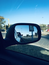 A car's side mirror reflects a visual of a street with other vehicles lined up, trees and a road sign in the distance. The sky is clear with a few clouds visible.