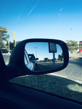 A car's side mirror reflects a visual of a street with other vehicles lined up, trees and a road sign in the distance. The sky is clear with a few clouds visible.