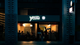 A dimly lit restaurant with a glowing sign that reads 'yuan.' The warm interior lighting contrasts with the darkness outside, showing a cozy dining area with several people seated around tables. Large windows reveal the scene inside, including hanging ceiling lights and a decorative plant.