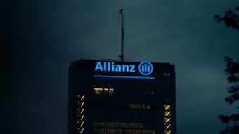 The top of a high-rise building is illuminated with blue light, showcasing the Allianz logo prominently. The surrounding sky is dark, creating a stark contrast with the building's lights.