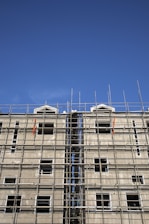 A multi-story building under construction with extensive scaffolding covering its facade. The scaffolding is composed of metal bars forming a grid-like structure, and the unfinished building has several window openings without glass. The sky above is clear and blue.