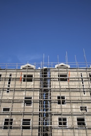 A multi-story building under construction with extensive scaffolding covering its facade. The scaffolding is composed of metal bars forming a grid-like structure, and the unfinished building has several window openings without glass. The sky above is clear and blue.