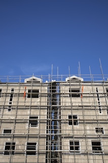 A multi-story building under construction with extensive scaffolding covering its facade. The scaffolding is composed of metal bars forming a grid-like structure, and the unfinished building has several window openings without glass. The sky above is clear and blue.