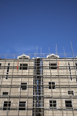 A multi-story building under construction with extensive scaffolding covering its facade. The scaffolding is composed of metal bars forming a grid-like structure, and the unfinished building has several window openings without glass. The sky above is clear and blue.