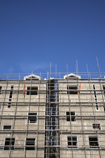 A multi-story building under construction with extensive scaffolding covering its facade. The scaffolding is composed of metal bars forming a grid-like structure, and the unfinished building has several window openings without glass. The sky above is clear and blue.