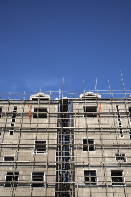 A multi-story building under construction with extensive scaffolding covering its facade. The scaffolding is composed of metal bars forming a grid-like structure, and the unfinished building has several window openings without glass. The sky above is clear and blue.