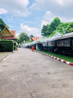 A peaceful outdoor scene featuring a solitary figure in an orange robe walking along a pathway, surrounded by lush greenery and structures with traditional architectural elements. There is a trail of mist or smoke along the path, and a bright blue sky with clouds overhead.