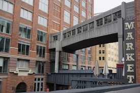 A large brick building with multiple windows, featuring an enclosed pedestrian walkway connecting to another structure. A sign with the word 'MARKET' is prominently displayed. The scene also includes closed patio umbrellas and a hint of another building in the background.