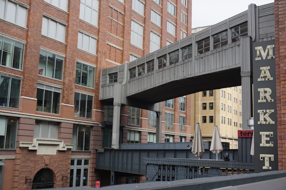 A large brick building with multiple windows, featuring an enclosed pedestrian walkway connecting to another structure. A sign with the word 'MARKET' is prominently displayed. The scene also includes closed patio umbrellas and a hint of another building in the background.