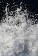 A dynamic and energetic splash of water captured mid-action with droplets and foam suspended in the air against a dark background. The movement creates a sense of power and fluidity.