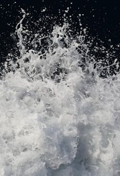 A dynamic and energetic splash of water captured mid-action with droplets and foam suspended in the air against a dark background. The movement creates a sense of power and fluidity.