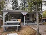 A small, rustic outdoor structure with a wooden roof and an underlying camper trailer. There is a picnic bench in the foreground and a weathered wooden path leading to the entrance. The setting is surrounded by tall trees, and a parked orange vehicle is partially visible on the right. Decorative elements include potted flowers and a covered patio with several chairs.