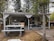 A small, rustic outdoor structure with a wooden roof and an underlying camper trailer. There is a picnic bench in the foreground and a weathered wooden path leading to the entrance. The setting is surrounded by tall trees, and a parked orange vehicle is partially visible on the right. Decorative elements include potted flowers and a covered patio with several chairs.