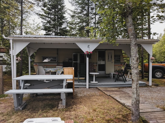 A small, rustic outdoor structure with a wooden roof and an underlying camper trailer. There is a picnic bench in the foreground and a weathered wooden path leading to the entrance. The setting is surrounded by tall trees, and a parked orange vehicle is partially visible on the right. Decorative elements include potted flowers and a covered patio with several chairs.
