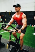 A man with a tattoo on his left arm is focused while using a stationary exercise bike. He is wearing a red tank top, camouflage shorts, and a black cap in a gym environment with artificial turf and gym equipment visible in the background.