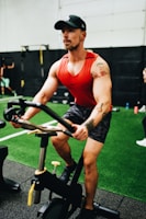 A man with a tattoo on his left arm is focused while using a stationary exercise bike. He is wearing a red tank top, camouflage shorts, and a black cap in a gym environment with artificial turf and gym equipment visible in the background.