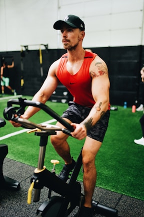 A man with a tattoo on his left arm is focused while using a stationary exercise bike. He is wearing a red tank top, camouflage shorts, and a black cap in a gym environment with artificial turf and gym equipment visible in the background.