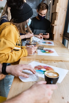 A group of people sitting at a wooden table engaged in an activity involving colorful charts. One person is holding a black coffee cup, while another person is drawing on a chart with a pencil. They are dressed casually, and some are wearing face masks, indicating it might be during the COVID-19 pandemic. The background includes some wood paneling and a window.